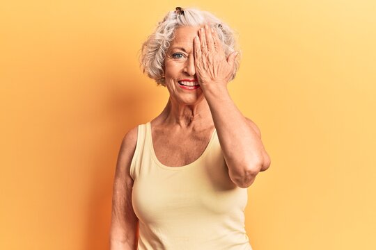 Senior grey-haired woman wearing casual clothes covering one eye with hand, confident smile on face and surprise emotion.