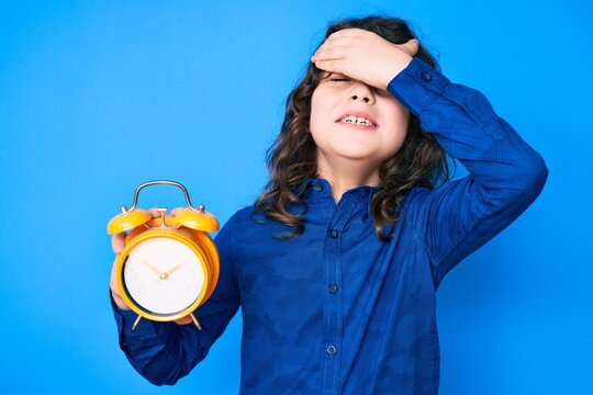 Cute hispanic child with long hair holding alarm clock stressed and frustrated with hand on head, surprised and angry face