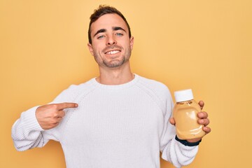 Young handsome man holding bottle of mustard sauce condiment over yellow background with surprise face pointing finger to himself