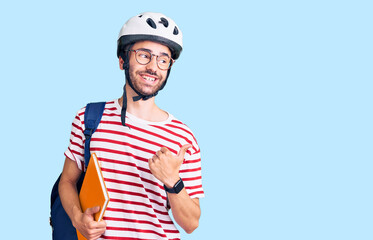 Young hispanic man wearing student backpack and bike helmet holding binder pointing thumb up to the side smiling happy with open mouth