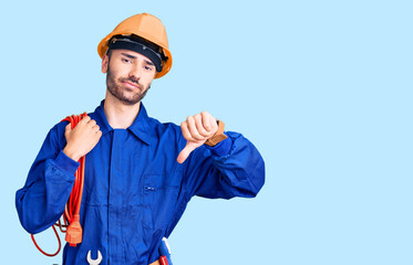 Young hispanic man wearing elecrician uniform holding cable with angry face, negative sign showing dislike with thumbs down, rejection concept