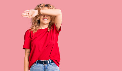 Young blonde woman with curly hair wearing casual red tshirt covering eyes with arm smiling...