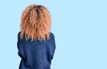 Young blonde woman with curly hair wearing casual winter sweater standing backwards looking away...