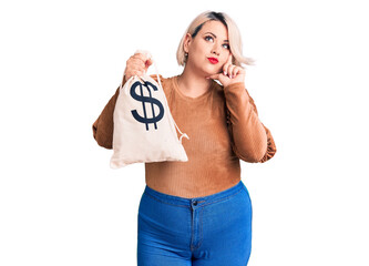 Young blonde plus size woman holding money bag with dollar symbol serious face thinking about question with hand on chin, thoughtful about confusing idea