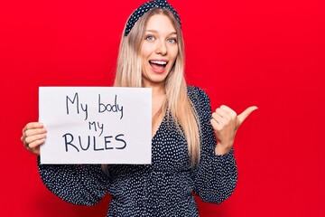 Young blonde woman holding my body my rules banner pointing thumb up to the side smiling happy with...