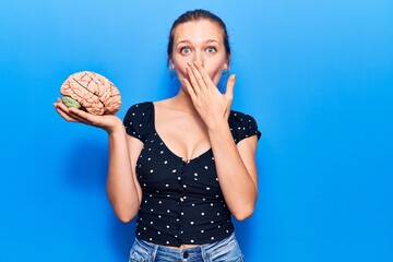 Young beautiful blonde woman holding brain covering mouth with hand, shocked and afraid for...