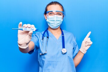 Young blonde woman wearing doctor uniform and coronavirus protection holding help reminder smiling happy pointing with hand and finger to the side