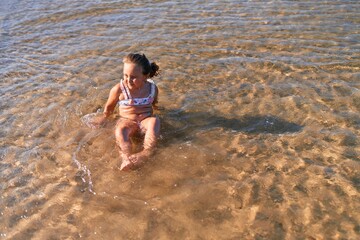 Adorable blonde child wearing bikini smiling happy. Sitting on the sand playing with water at the sea