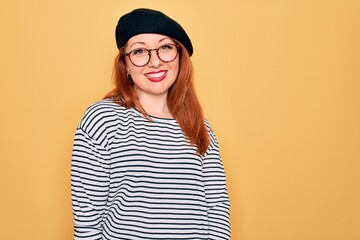 Beautiful redhead woman wearing striped t-shirt and french beret over yellow background with a happy and cool smile on face. Lucky person.