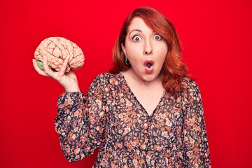 Young beautiful redhead woman asking for care memory holding brain over red backgroud scared and...