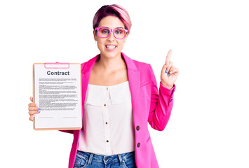 Young beautiful woman with pink hair holding clipboard with contract document with a big smile on face, pointing with hand finger to the side looking at the camera.