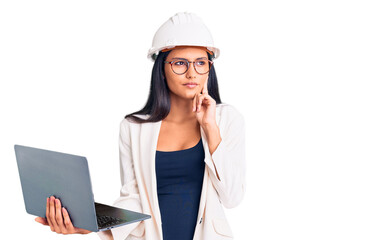 Young beautiful latin girl wearing architect hardhat holding laptop serious face thinking about question with hand on chin, thoughtful about confusing idea