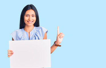 Fototapeta premium Young beautiful latin girl holding blank empty banner surprised with an idea or question pointing finger with happy face, number one