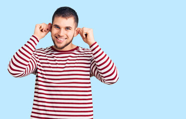 Young handsome man wearing striped sweater smiling pulling ears with fingers, funny gesture....