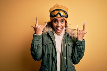 Young african american afro skier girl wearing snow sportswear and ski goggles shouting with crazy expression doing rock symbol with hands up. Music star. Heavy music concept.