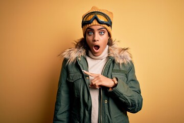 Young african american afro skier girl wearing snow sportswear and ski goggles Surprised pointing with finger to the side, open mouth amazed expression.