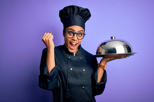 Young african american cooker girl wearing uniform and hat holding tray with dome screaming proud and celebrating victory and success very excited, cheering emotion