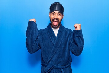 Naklejka premium Young hispanic man wearing sleep mask and robe screaming proud, celebrating victory and success very excited with raised arms