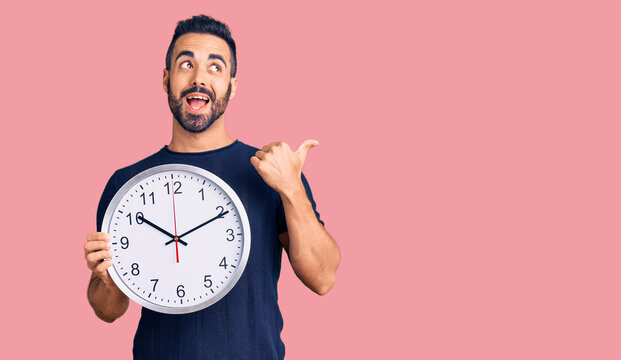 Young hispanic man holding big clock pointing thumb up to the side smiling happy with open mouth