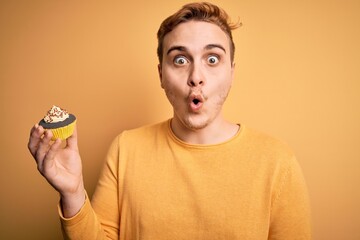 Young handsome redhead man eating sweet chocolate cupcake dessert over yellow background scared and...