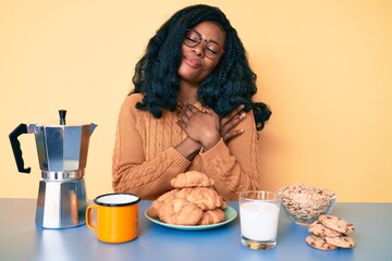 Beautiful african woman sitting on the table eating breakfast in the morning smiling with hands on...
