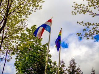 LGBT and Holland flags on poles blow in the wind. Symbol of tolerant. Gay sign rainbow. The rainbow pride, Trees and blue cloudy sky on the background. Copy space