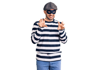 Young handsome man wearing burglar mask smiling funny doing claw gesture as cat, aggressive and...