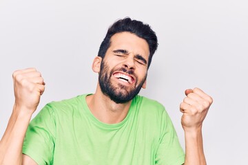 Young handsome man with beard wearing casual t-shirt very happy and excited doing winner gesture with arms raised, smiling and screaming for success. celebration concept.