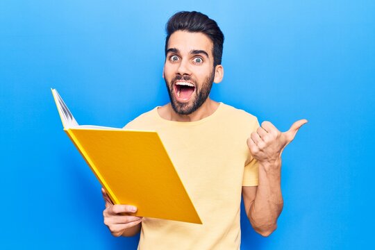 Young handsome man with beard reading book pointing thumb up to the side smiling happy with open mouth