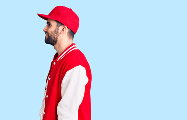 Young handsome man with beard wearing baseball jacket and cap looking to side, relax profile pose with natural face with confident smile.