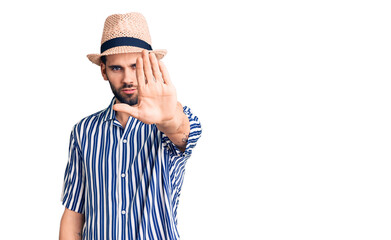 Young handsome man with beard wearing summer hat and striped shirt doing stop sing with palm of the hand. warning expression with negative and serious gesture on the face.