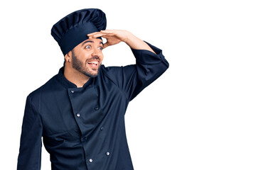 Young hispanic man wearing cooker uniform very happy and smiling looking far away with hand over head. searching concept.