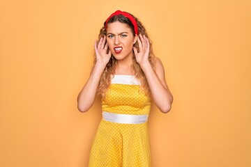 Beautiful blonde pin-up woman with blue eyes wearing diadem standing over yellow background Trying to hear both hands on ear gesture, curious for gossip. Hearing problem, deaf