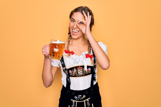Beautiful blonde german woman with blue eyes wearing octoberfest dress drinking jar of beer with happy face smiling doing ok sign with hand on eye looking through fingers