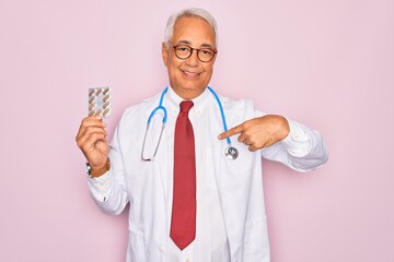 Middle age senior grey-haired doctor man holding prescription pills over pink background with surprise face pointing finger to himself