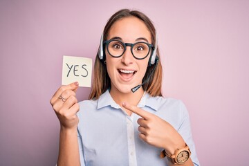 Beautiful call center agent woman working using headset holding reminder with yes message very...