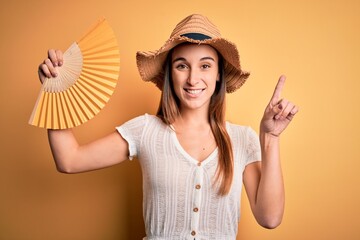 Young tourist woman on vacation wearing summer hat holding hand fan over yellow background surprised with an idea or question pointing finger with happy face, number one