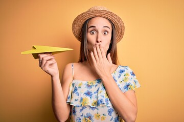 Young tourist woman on vacation wearing summer hat holding hand fan over yellow background cover mouth with hand shocked with shame for mistake, expression of fear, scared in silence, secret concept