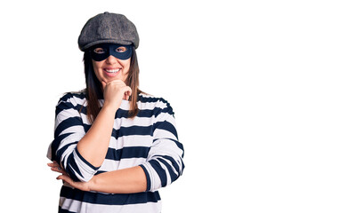 Young beautiful brunette woman wearing burglar mask looking confident at the camera smiling with crossed arms and hand raised on chin. thinking positive.