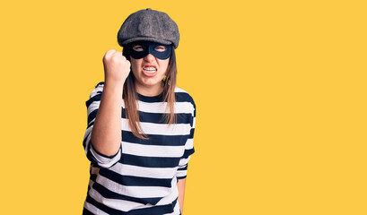 Young beautiful brunette woman wearing burglar mask angry and mad raising fist frustrated and furious while shouting with anger. rage and aggressive concept.