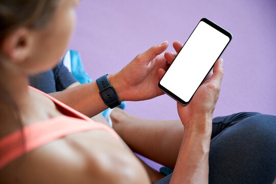 Over shoulder closeup view of fit sporty woman yoga teacher sit on mat holding phone mock up screen video call in zoom, watching online workout training class video, using fitness app at home in gym.