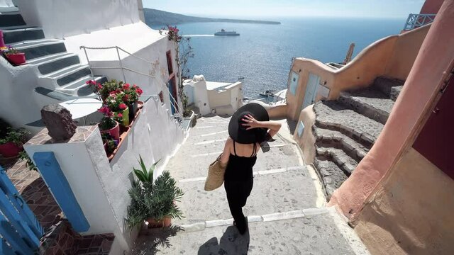 Tourist travel woman in Oia, Santorini, Greece. Happy elegant fashion model in black walking on stairs by famous landmark destination in Greece Santorini