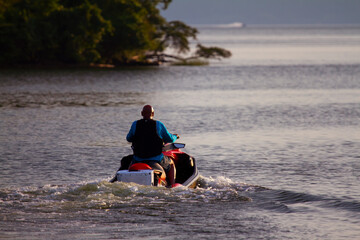 Fototapeta na wymiar A man with a shaved head wearing life vest is riding a personal water craft in mallows bay of Potomac river at sunset. There are trees in the blurred background.