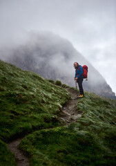 Man traveler with backpack standing on grassy hillside path with foggy mountain on background. Handsome tourist looking at camera while hiking in mountains. Concept of traveling and climbing.