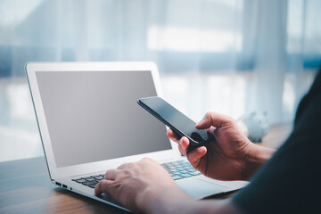 Businessman's hand is typing a keyboard on a laptop and holding a cellphone with a blank screen. Close-up of Man's hands working on desk.