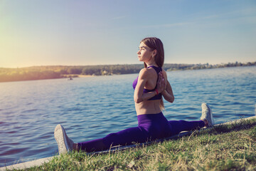 young european female athlete doing exercises for stretching the legs, sits on the splits outside, near the lake. Healthy lifestyle.
