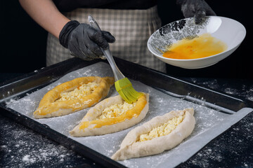 Woman in checkered apron and black gloves covers raw bakery khachapuri with egg yolk. Adjarian Khachapuri Recipe – Georgian cheese bread.