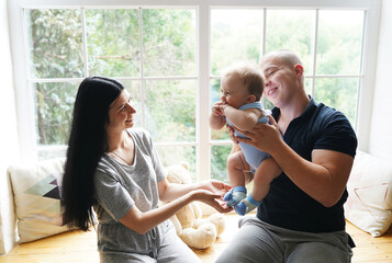 Mother, father and their baby son having fun and playing at home near a big window with sunlight