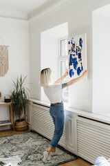 Blonde caucasian woman placing abstract black and phantom blue paint on the wall near the window...