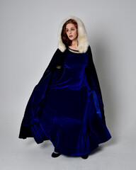 Full length portrait of  girl wearing long blue velvet gown with golden crown. sitting pose, isolated against a studio background.